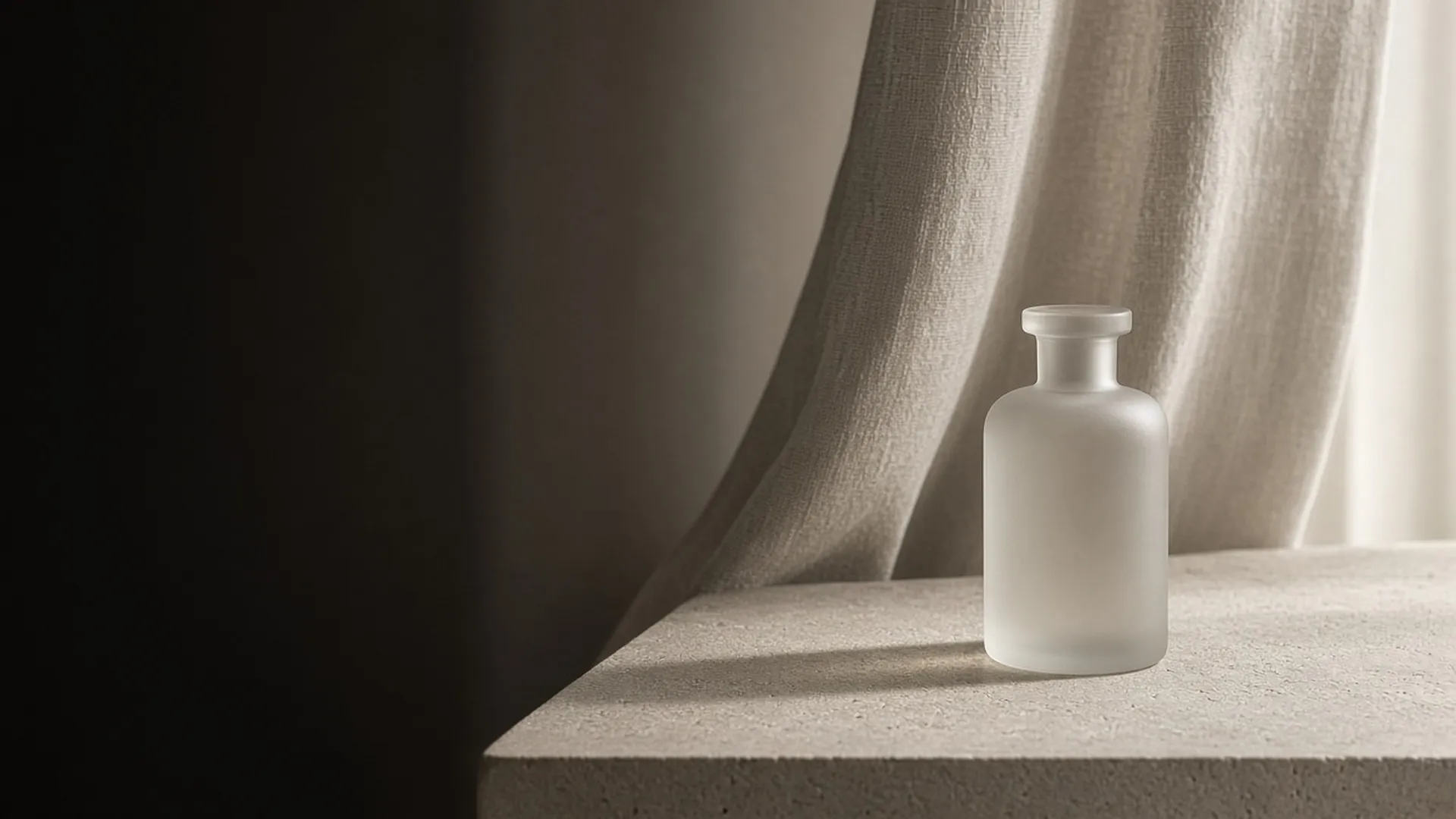 A plain frosted glass bottle on a stone ledge in front of a linen curtain