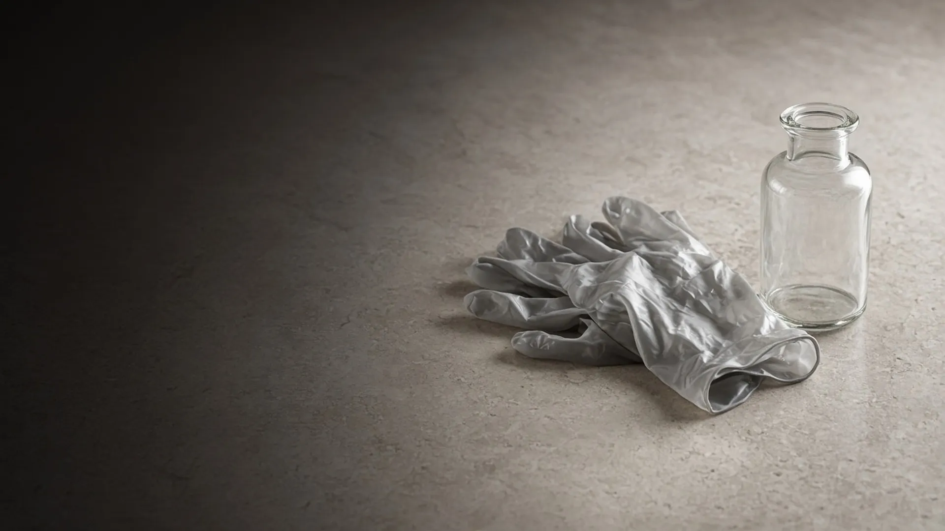 A pair of crumpled grey gloves beside a small clear unlabeled glass bottle on stone