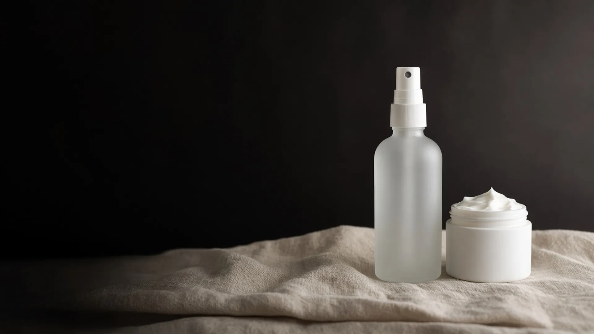 A plain frosted spray bottle beside a small open jar of white cream on linen