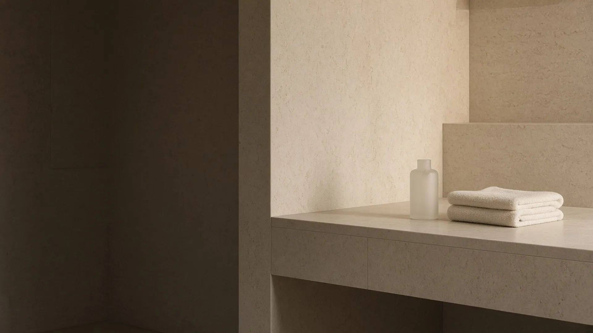 A single plain bottle and a folded towel on a limestone vanity in evening light