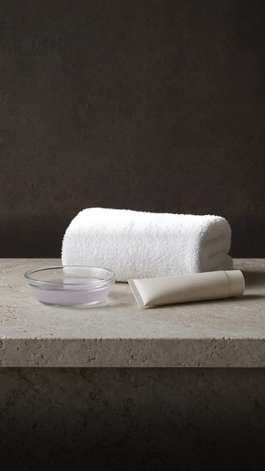 A white tube and rolled towel beside a clear unlabeled glass dish of pale lilac liquid