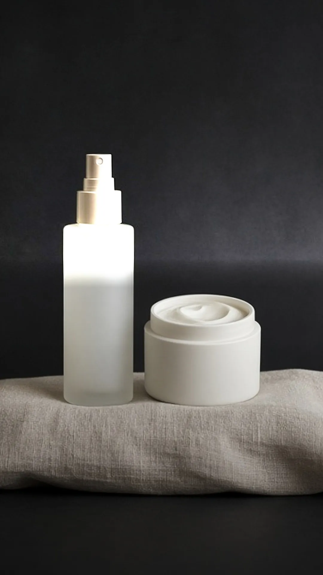 A plain frosted spray bottle beside a small open jar of white cream on linen