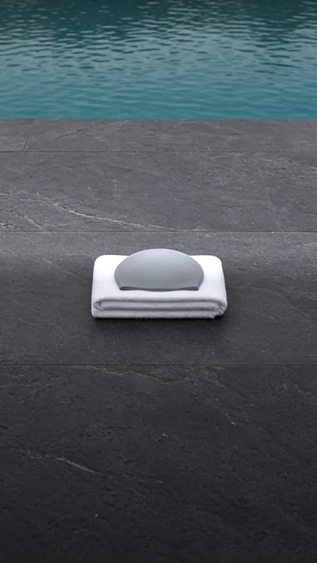 A folded white towel with a flat grey disc on a dark pool deck beside water